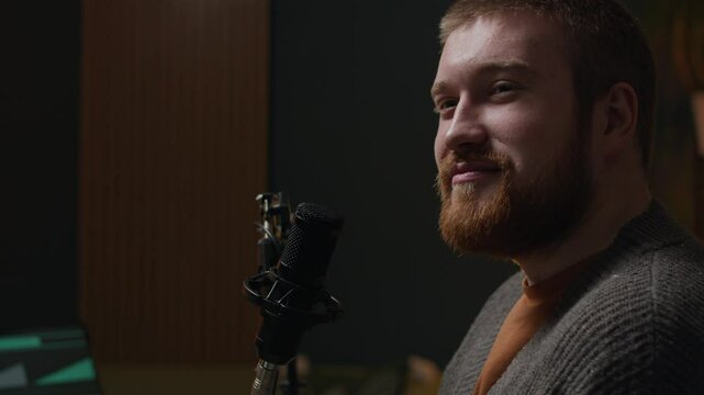 Close up shot of young Caucasian male radio host with beard laughing and answering question in mic sitting in modern broadcasting studio with acoustic panels and ring light in blurred background