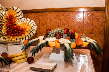 The image shows a table with a fruit arrangement. In the foreground is a tray with a variety of fruits such as grapes (green and red), strawberries, orange slices, pineapple and peaches