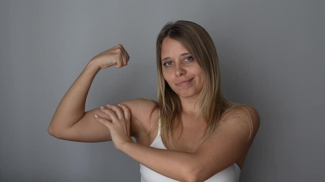 Young strong fit blonde woman raising arm and showing bicep grabbing muscles with her hand on a dark grey  background. Feminism, girl power, equal women's rights, independence, sports concept