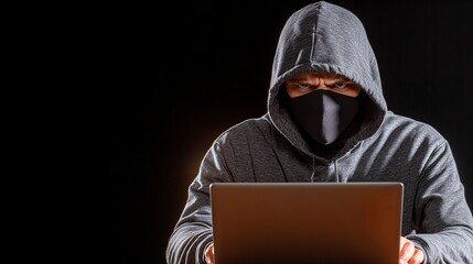 A hooded figure, face obscured by a mask, sits intently before a laptop in a dimly lit setting. The mysterious image evokes a sense of secrecy and intrigue.