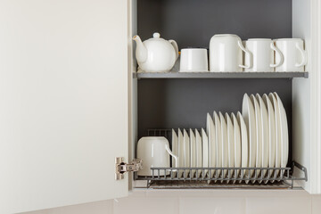 A dish-drying rack integrated into a wall cabinet in a modern kitchen with white ceramic kitchenware, plates and mugs, arranged orderly