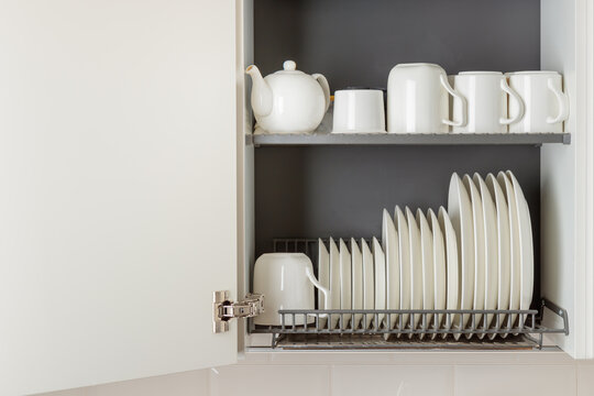 A dish-drying rack integrated into a wall cabinet in a modern kitchen with white ceramic kitchenware, plates and mugs, arranged orderly