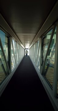 Walking through jetty bridge from landed airplane to modern airport terminal, first-person view, vertical slow motion shot. Typical telescopic corridor with glass walls, seems to be semi-empty flight