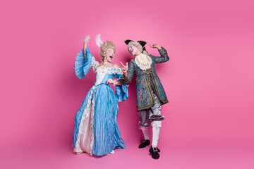 Elegant couple in baroque costumes enjoying a playful moment against a vibrant pink background, showcasing historical elegance