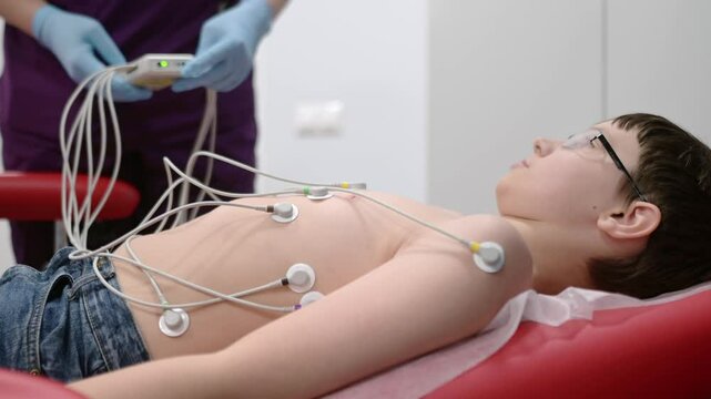 Child under ECG test with doctor. Boy patient lying in medical bed with vacuum sensors. A nurse performs an electrocardiogram of the heart in office of a medical clinic. Checkup of heart