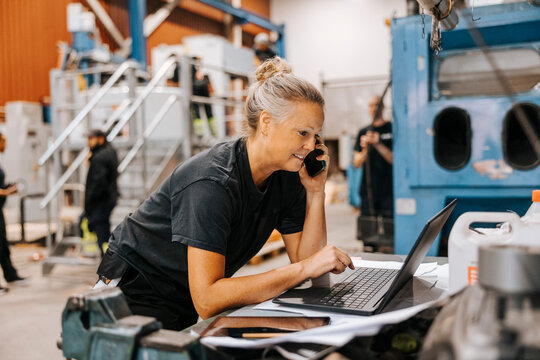 Female warehouse worker talking on smart phone while using laptop in manufacturing factory