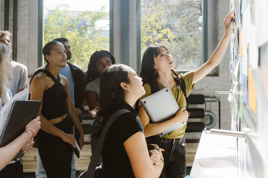 Young woman holding laptop while pointing at bulletin board while standing with university students in college
