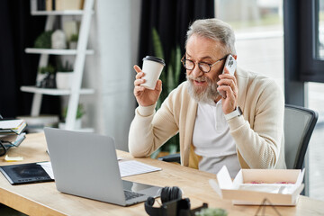 A mature handsome man is on a business call, holding a coffee cup while sitting at his desk.