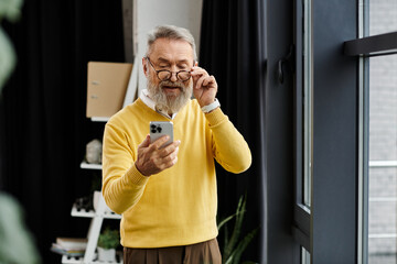 Mature man in a yellow sweater using his smartphone while standing in a stylish room