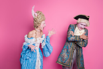 Elegant couple dressed in lavish baroque costumes stand against a vibrant pink backdrop, exuding historical charm