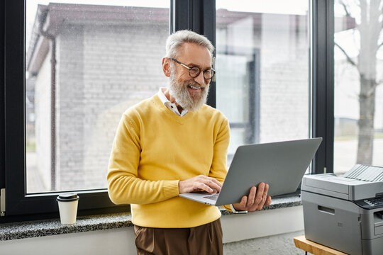 A mature handsome man smiles while using a laptop near a window in a bright office.
