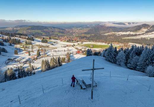 Woman on a winter hike, enoying the stunning view from the Imberg summit cross down to Steibis and Oberstaufen, Allgaeu Alps, Bavaria, Germany