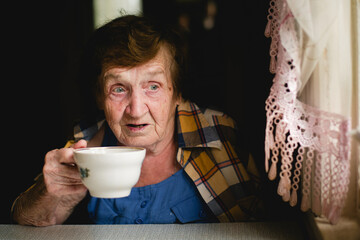 An elderly woman sits thoughtfully, sipping tea, lost in her own reflections.
