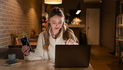 Young blonde woman working with her laptop and using charging with power bank, sitting in the cozy...