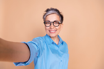Charming elderly woman wearing trendy blue striped shirt and glasses takes a joyful selfie against...