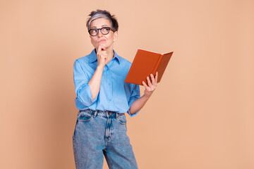 Thoughtful senior woman in casual attire holding a book and contemplating against a beige background