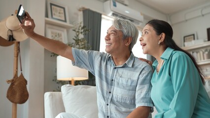 Obraz premium Happy grandfather and grandmother making video call with their son at home. Elder man holding smart phone and sitting at sofa while old woman greeting with cheerful vibe at modern house. Myrmidon.