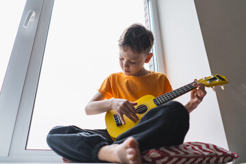 A boy dressed in an orange shirt focused on strumming a bright yellow ukulele in a cozy his room at...