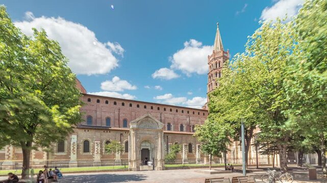 The cobbled parvis of the Basilica of Saint Sernin in Toulouse, France, timelapse hyperlapse. Largest romanesque church in Europe with brick bell tower, arched doors, green trees and blue cloudy sky