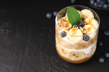 Tasty trifle dessert. Sponge cake, blueberries, banana and whipped cream in glass on black table, closeup. Space for text