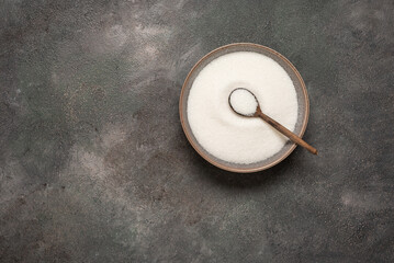 White sugar in bowl with wooden spoon on dark grunge background, top view