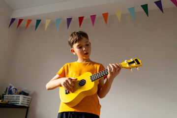 A boy dressed in an orange shirt focused on strumming a bright yellow ukulele in a cozy his room at...