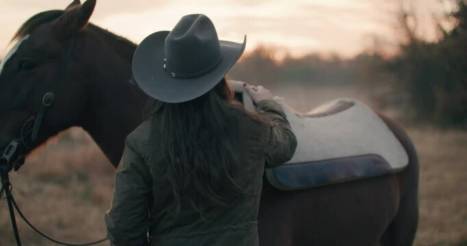 Woman Wearing Cowboy Hat Putting On and Adjusting Saddle On Horse In Grassy Meadow Surrounded By Trees At Sunset