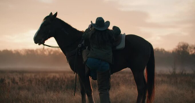 Cowgirl Woman Wearing Cowboy Hat Climbing Onto Stationary Horse and Riding Off Into the Sunset Through Meadow Surrounded By Trees 