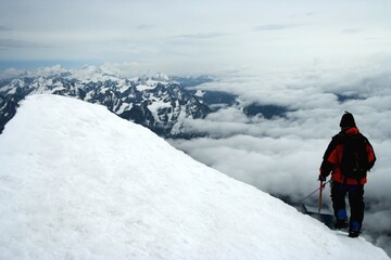 A climber ascends the snowy slopes of Huayna Potosí in Bolivia, surrounded by breathtaking mountain views and clouds.