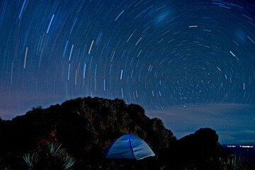 A long-exposure photograph captures star trails forming circular patterns in the night sky above a tent set up on rocky terrain in Bolivia. The image highlights the Earth's rotation.