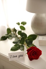 Paper with text I Love You and red rose on nightstand indoors