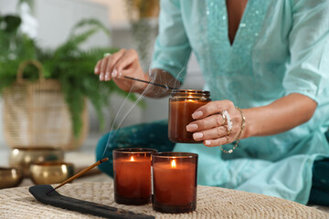 Woman lighting incense sticks with burning candles indoors, closeup