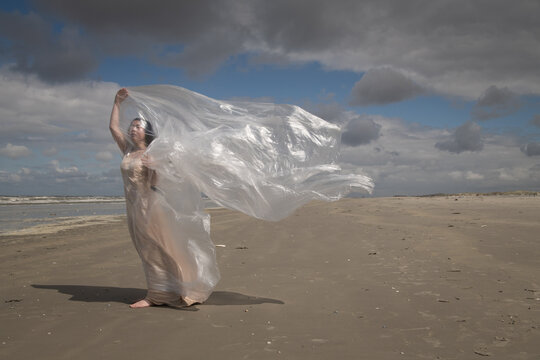 art portrait of woman in long dress standing on beach near ocean waving thin plastic sheet in the wind 