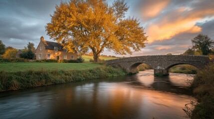 Obraz premium Idyllic autumn scene cottage, stone bridge, river, and golden tree at sunset.