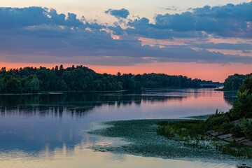 Serene river landscape at sunset with reflections of trees and sky, soft clouds, and distant bridge creating a peaceful, atmospheric view.  

