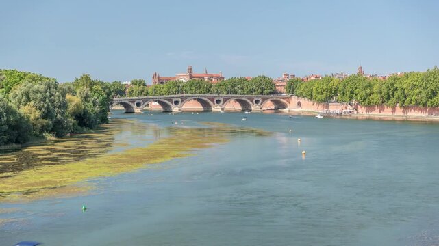 Garonne River and Pont Neuf timelapse in downtown Toulouse, France. Renaissance arch bridge reflects in the water under a blue sky with Basilica of Our Lady of the Daurade. Waterfront with green trees