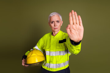 Confident female worker in high visibility uniform and helmet against khaki background demonstrates stop gesture