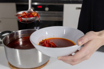 Serving hot borscht with vegetables into a white bowl from a pot in a home kitchen setting