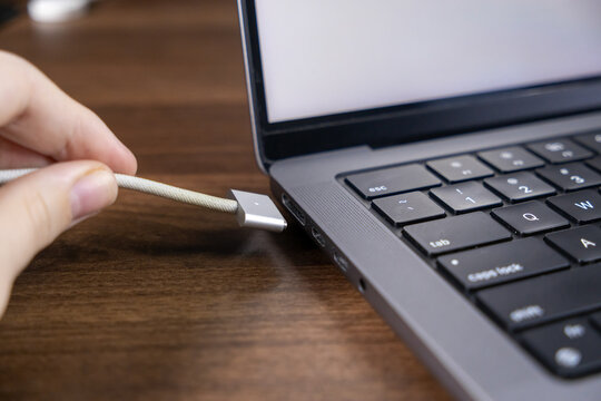 Hand plugging MagSafe charging cable into a modern laptop on a wooden desk