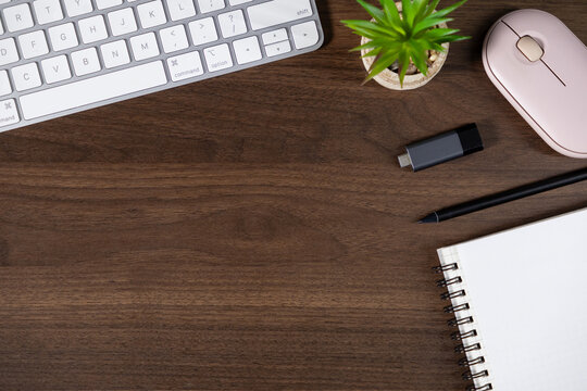 organized workspace with keyboard, notebook, pen, flash drive, plant, and wireless mouse on wooden desk. top view