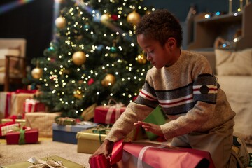 Side view portrait of little African American boy opening Christmas presents and ripping wrapping paper while celebrating holidays and enjoying surprise copy space