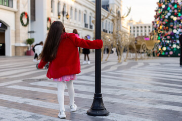 A girl in a red coat twirls around a lamppost in a Christmas square, surrounded by decorations and a giant Christmas tree.