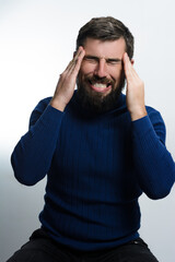 Fototapeta premium A man with a beard, wearing a blue sweater, grimaces in pain while holding his head. He appears to be suffering from a severe headache or migraine against a light background.