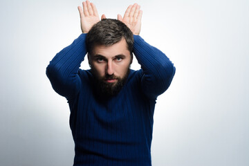 A bearded man in a blue sweater playfully gestures with his hands above his head, forming bunny ears against a light gray background.