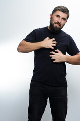 A man with a beard, wearing a black shirt and jeans, scratches his chest with both hands. He...