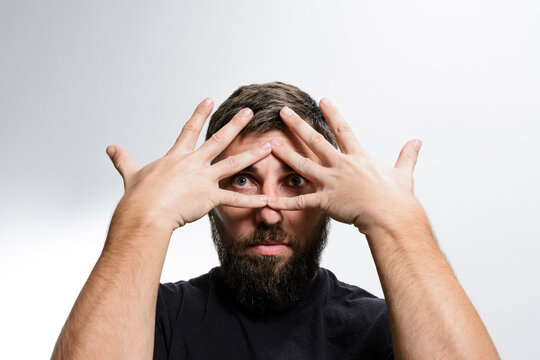 A bearded man looks intently at the camera through a triangular opening formed by his fingers, conveying curiosity, scrutiny, or playful observation.