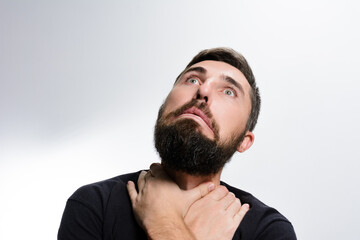 Obraz premium A bearded man against a light background looks upward with wide eyes and a worried expression. His hands are clasped around his neck, suggesting concern or discomfort.