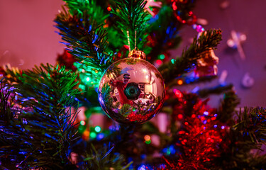 Reflection of a photographer in a shiny reddish ball ornament hanging on a Christmas tree with LED lights and colorful garlands.