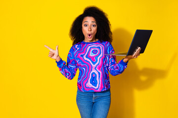 Photo of attractive young woman hold netbook amazed point empty space dressed stylish colorful blue clothes isolated on yellow background