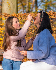 Fototapeta premium Mother and cute preschool daughter have fun together in nature in fall park, paint lips with lipstick, do each other's makeup, have nice time at home.Happy family.